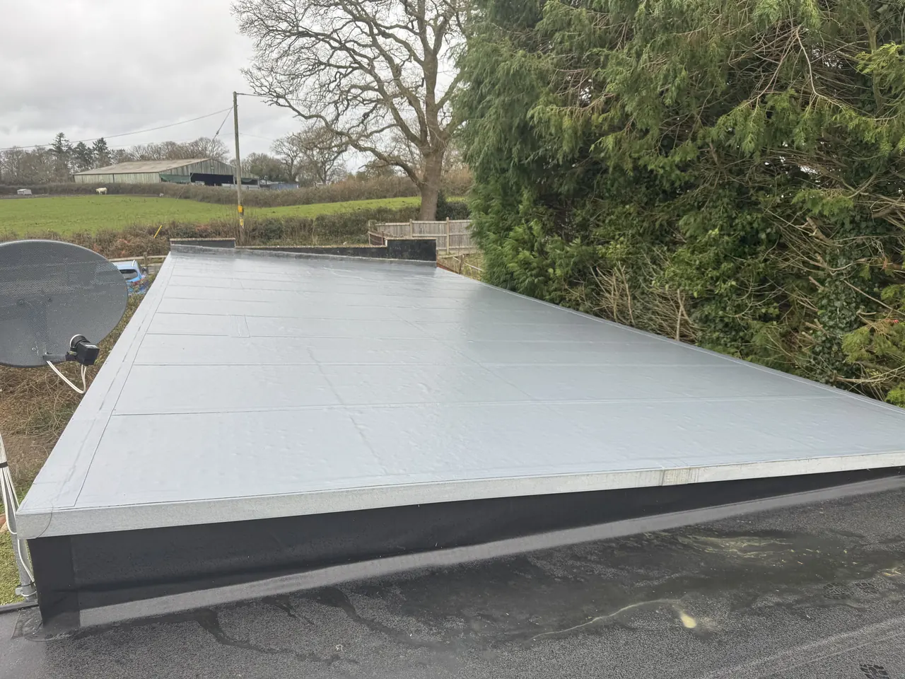 Large light grey fibreglass flat roof with dark fascia, fields and a barn behind
